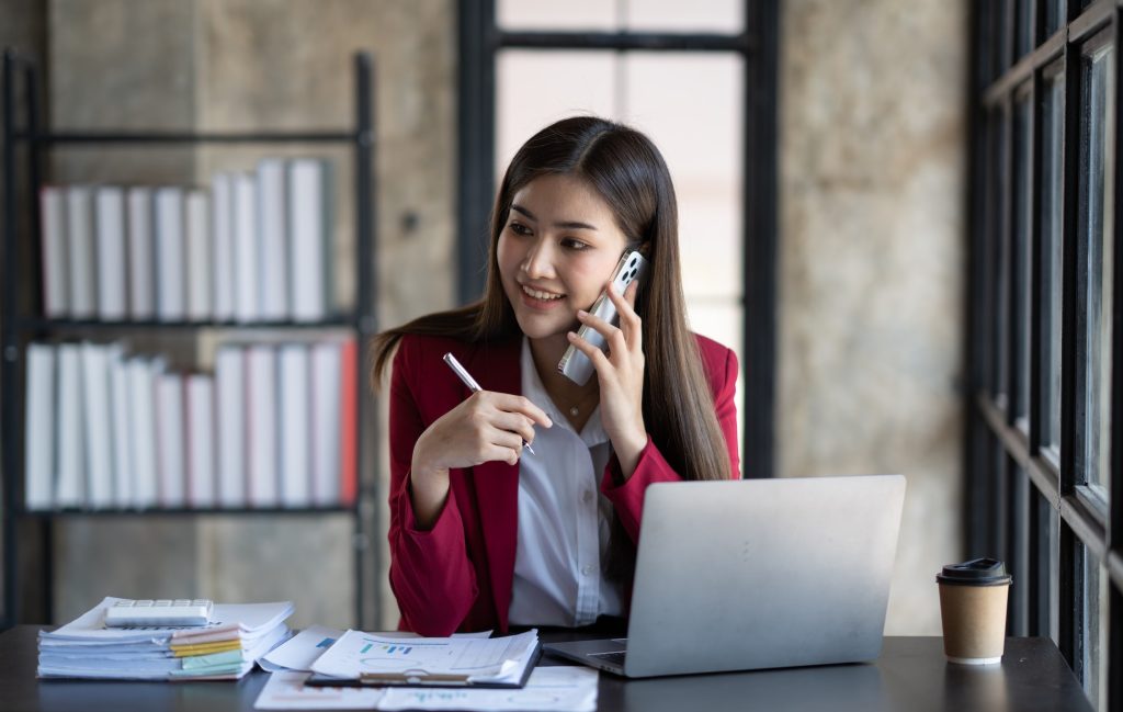 Happy businesswoman using mobile phone while working at office with laptop. Smiling woman contact cu